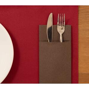 Formal place setting: knife and fork tucked into a brown napkin pocket on a red placemat, with a white plate and wood table edge visible.